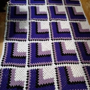 May include: A crocheted blanket with a geometric pattern in shades of purple, black, and white.