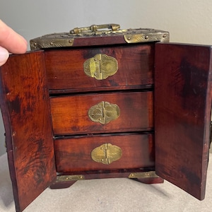 May include: A small, open wooden jewelry box with three drawers. The box is a dark reddish-brown color with brass hardware, including decorative clasps and corner accents. The doors are open, revealing the drawers.