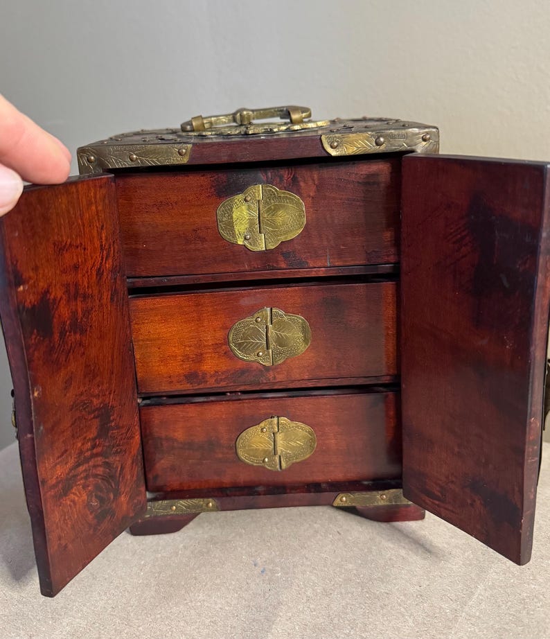 May include: A small, open wooden jewelry box with three drawers. The box is a dark reddish-brown color with brass hardware, including decorative clasps and corner accents. The doors are open, revealing the drawers.
