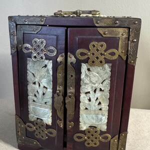 May include: A small, dark red wooden cabinet with two doors, adorned with ornate brass hardware and corner brackets. The doors feature carved white panels with floral designs, and decorative brass clasps.