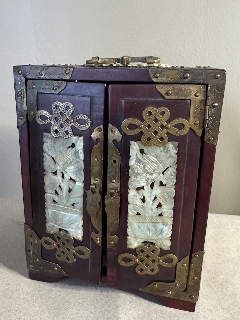 May include: A small, dark red wooden cabinet with two doors, adorned with ornate brass hardware and corner brackets. The doors feature carved white panels with floral designs, and decorative brass clasps.