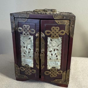 May include: A small, dark red wooden cabinet with brass hardware and decorative corner brackets. The cabinet doors feature carved white jade panels with bird and floral designs, and ornate metal accents. A handle is on top.