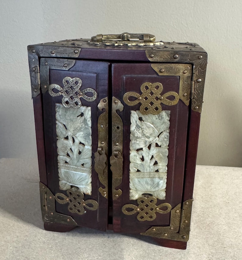 May include: A small, dark red wooden cabinet with brass hardware and decorative corner brackets. The cabinet doors feature carved white jade panels with bird and floral designs, and ornate metal accents. A handle is on top.