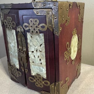 May include: A small, antique wooden cabinet with a dark red finish. The cabinet features ornate brass corner protectors and decorative jade panels with carved bird and floral designs. The doors are secured with brass hardware.