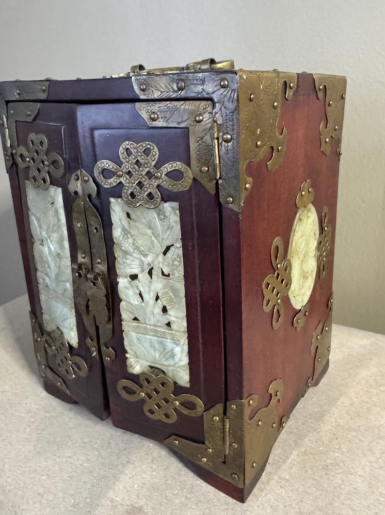 May include: A small, antique wooden cabinet with a dark red finish. The cabinet features ornate brass corner protectors and decorative jade panels with carved bird and floral designs. The doors are secured with brass hardware.