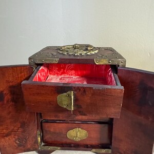 May include: A wooden jewelry box with two drawers and a top compartment. The box is dark brown with brass accents on the corners, handle, and clasps. The interior is lined with red fabric.