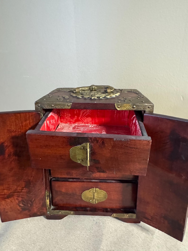May include: A wooden jewelry box with two drawers and a top compartment. The box is dark brown with brass accents on the corners, handle, and clasps. The interior is lined with red fabric.