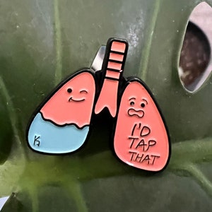 May include: A pink and blue enamel pin shaped like a pair of lungs. The left lung has a smiling face and the right lung has a surprised face and says "I'd Tap That".
