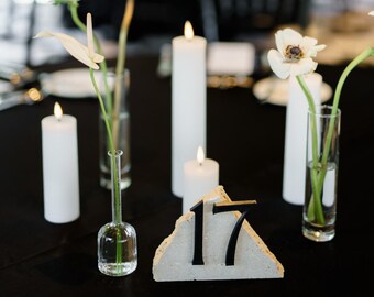 May include: A table setting with white candles, clear glass vases holding white flowers and greenery, and a concrete table number 17 with black numerals. The table is covered with a black tablecloth.