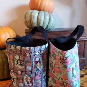 May include: Two Halloween trick-or-treat bags with black handles. One bag features a purple and pink stained glass pattern with images of Jack Skellington and Sally from The Nightmare Before Christmas. The other bag has a green background with pink roses and images of Jack Skellington and Sally.