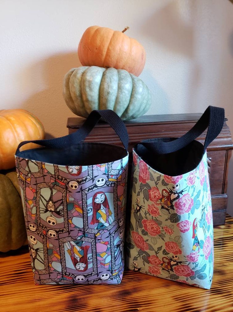 May include: Two Halloween trick-or-treat bags with black handles. One bag features a purple and pink stained glass pattern with images of Jack Skellington and Sally from The Nightmare Before Christmas. The other bag has a green background with pink roses and images of Jack Skellington and Sally.