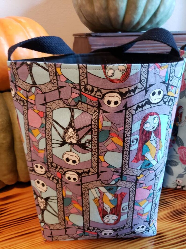 May include: A black canvas tote bag with a Nightmare Before Christmas print. The print features Jack Skellington and Sally in a stained glass style. The bag has black handles.