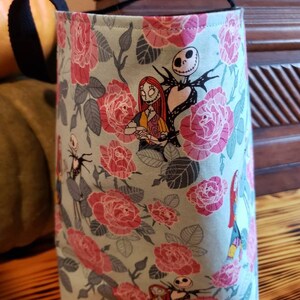 May include: A teal fabric tote bag with black handles featuring a floral pattern of pink roses and leaves. The bag has a print of Jack Skellington and Sally from the Nightmare Before Christmas.
