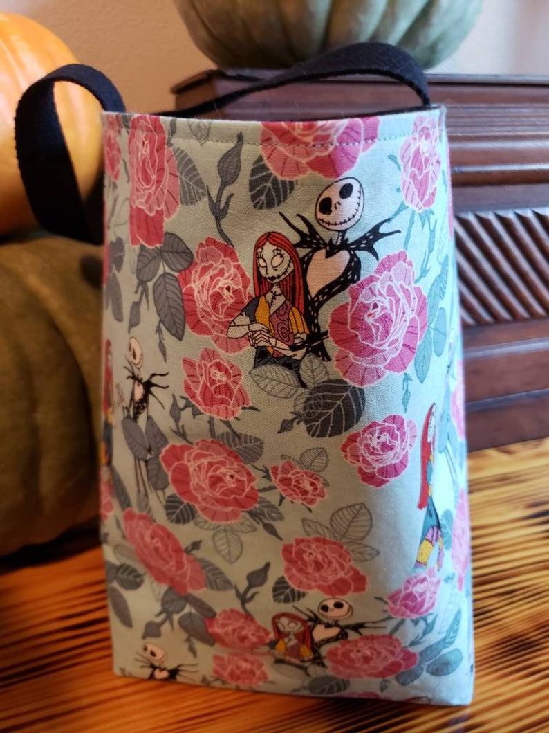 May include: A teal fabric tote bag with black handles featuring a floral pattern of pink roses and leaves. The bag has a print of Jack Skellington and Sally from the Nightmare Before Christmas.