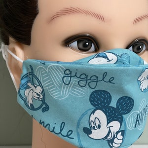 May include: A light blue face mask featuring a repeating pattern of Mickey Mouse and other cartoon characters. The mask has the words "giggle" and "smile" printed on it. The mask is made of fabric and has white elastic ear loops.