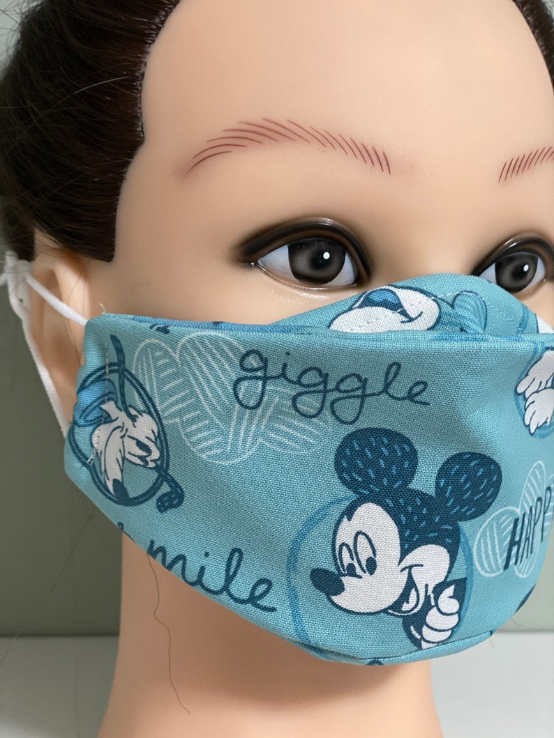 May include: A light blue face mask featuring a repeating pattern of Mickey Mouse and other cartoon characters. The mask has the words "giggle" and "smile" printed on it. The mask is made of fabric and has white elastic ear loops.