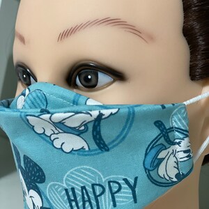 May include: A light blue face mask with a repeating pattern of a cartoon character and the word "HAPPY" in white. The mask has white elastic ear loops.
