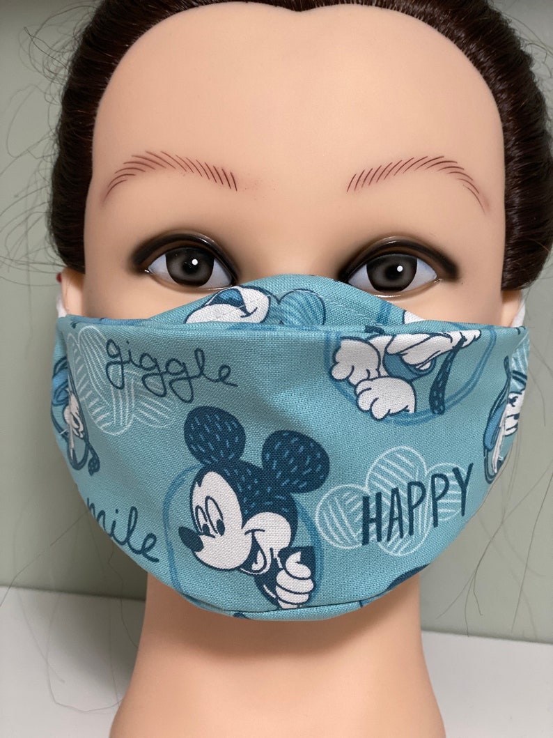 May include: A light blue face mask featuring a repeating pattern of Mickey Mouse illustrations and the words "giggle", "smile", and "happy". The mask has white elastic ear loops.