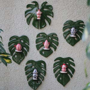 May include: A collection of ceramic wall decorations featuring green monstera leaves with small decorative vases. The vases have various designs, including stripes and floral patterns, and are in shades of red, white, and blue.