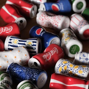 May include: A collection of ceramic cups shaped like crushed beverage cans, featuring various designs including Coca-Cola, Pepsi, and Heineken logos, alongside floral, lemon, and striped patterns in red, blue, green, and white colour schemes.