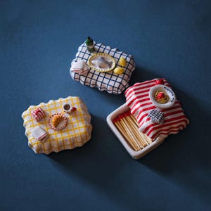 Puede incluir: Tres cajas de cerillas rectangulares de cerámica con tapas decorativas. Una tiene un patrón de cuadrícula azul con un pescado, otra una cuadrícula amarilla con un cruasán y otra un patrón de rayas rojas con un plato de pasta con la palabra mamma mia.
