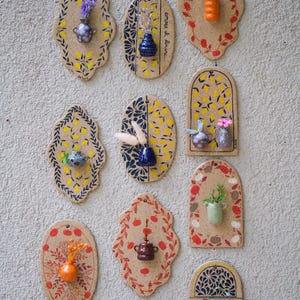 May include: A collection of ceramic wall art pieces, each with a unique shape and design. The pieces feature small vases with flowers and decorative patterns in yellow, blue, and red. The art is mounted on a textured, light-colored wall.