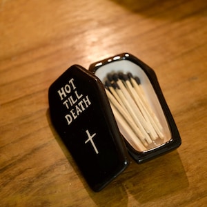 Peut inclure: Un porte-allumettes noir en forme de cercueil avec le texte "HOT TILL DEATH" et une croix. Le support est ouvert, révélant des allumettes en bois à bouts noirs. Le support est sur une surface en bois.