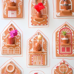 Può includere: Una collezione di case decorative da parete color terracotta, ognuna con un design unico e dettagli bianchi. Alcune case hanno piccoli vasi con fiori, altre ghirlande o elementi decorativi, creando un'esposizione affascinante e stravagante.