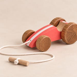 May include: A wooden pull toy shaped like a race car, featuring a smooth red body with a white center stripe, four natural wood-grain wheels, and a white cotton pull cord attached to a small cylindrical wooden handle for easy gripping.