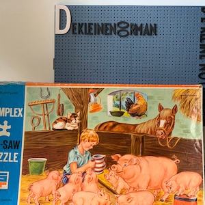 May include: A vintage children's jigsaw puzzle box with a blue and white design. The box features a picture of a boy feeding piglets in a barn. The text on the box reads "SIMPLEX IG-SAW PUZZLE 26 delen/pieces".