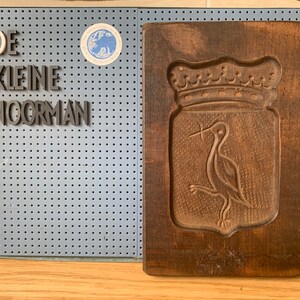May include: A wooden cookie cutter with a carved design of a bird with a crown above it. The cutter is dark brown and has a rectangular shape. The text "DE KLEINE NOORMAN" is visible on a blue pegboard behind the cutter.