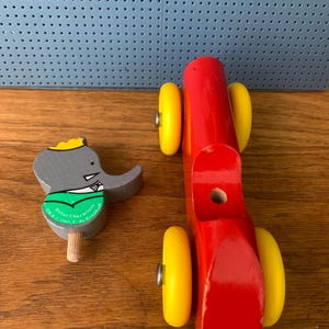 May include: A wooden toy set consisting of a bright red vehicle body with four yellow wheels and a separate gray elephant figure wearing a green shirt and yellow crown, featuring the text Babar Characters TM & C 1989 L. de Brunhoff.