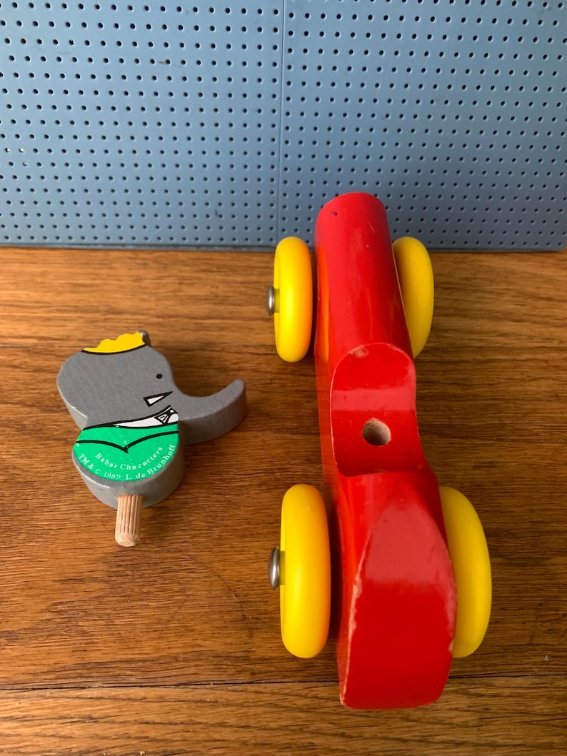 May include: A wooden toy set consisting of a bright red vehicle body with four yellow wheels and a separate gray elephant figure wearing a green shirt and yellow crown, featuring the text Babar Characters TM & C 1989 L. de Brunhoff.