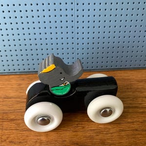 May include: A wooden toy car featuring a black body and four white wheels with silver centers. A gray elephant figure with a yellow ear and a green shirt is attached to the top. The elephant has a small white triangle detail on its chest.