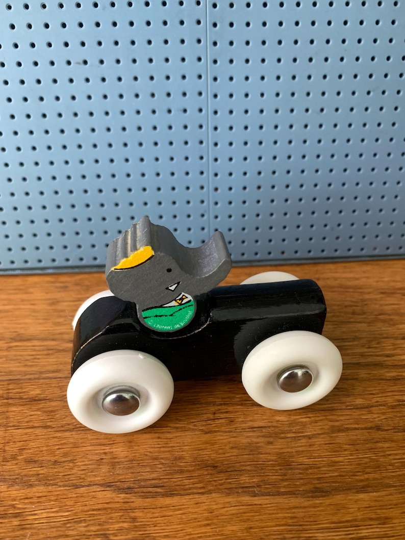 May include: A wooden toy car featuring a black body and four white wheels with silver centers. A gray elephant figure with a yellow ear and a green shirt is attached to the top. The elephant has a small white triangle detail on its chest.