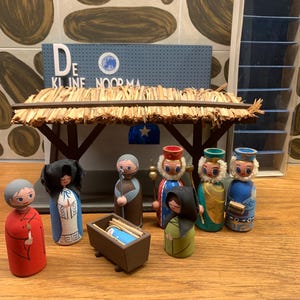 May include: A vintage wooden nativity scene with figures and a stable. The figures are painted in bright colors, including red, blue, and green. The stable has a thatched roof and a blue sign that reads "De KLINE NOORMA".