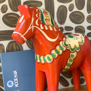 Op de afbeelding: Een felrood, handgeschilderd houten Dala-paard, versierd met witte, groene en gele bloempatronen. Het paard heeft een gedetailleerde manen- en zadelontwerp. Een blauw boek met witte tekst staat op de achtergrond.