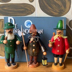 May include: Three wooden figurines with detailed features. The first is green with a white beard and hat, holding a pipe and rifle. The second is brown with a black cape, holding a spear and lantern. The third is red with a green hat, holding a staff.