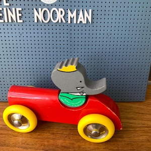 May include: A wooden toy car painted bright red with four yellow wheels and silver hubcaps. A gray elephant character with a yellow crown and green shirt is seated in the driver's position. The figure features text reading TM & &copy; 1990 L. de Brunhoff.
