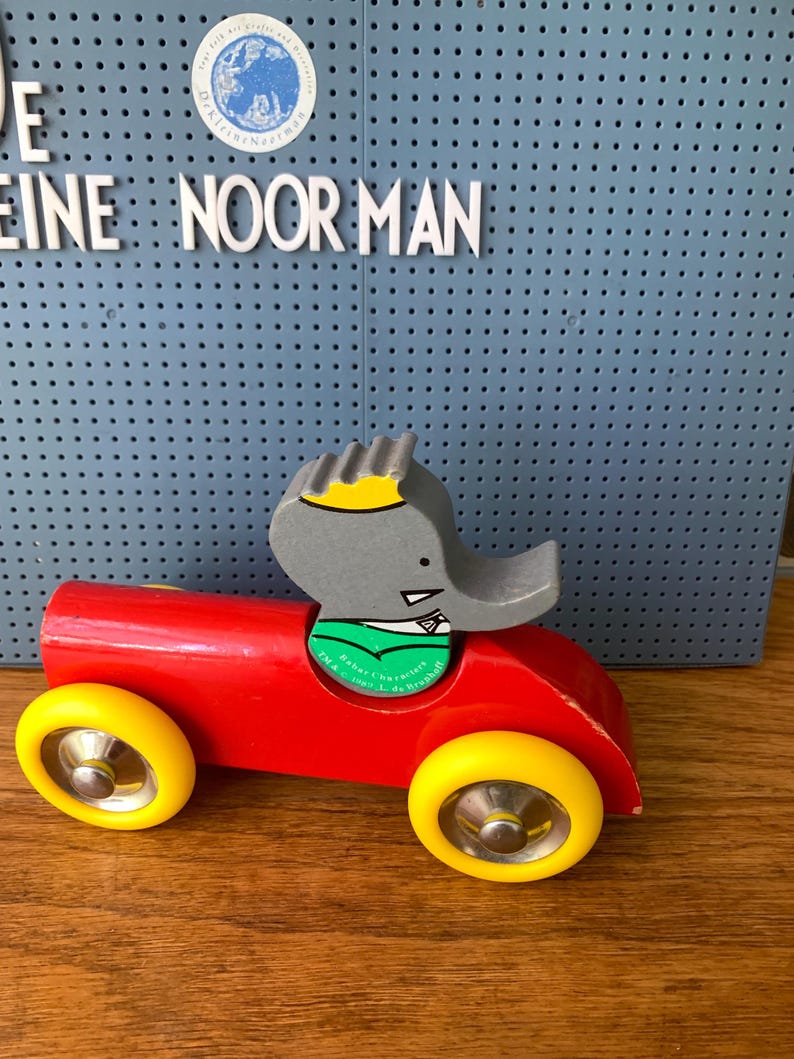 May include: A wooden toy car painted bright red with four yellow wheels and silver hubcaps. A gray elephant character with a yellow crown and green shirt is seated in the driver's position. The figure features text reading TM & &copy; 1990 L. de Brunhoff.