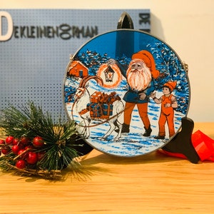 May include: A round, blue and white Christmas plate with a Santa Claus figure carrying a lantern and a goat carrying presents. The plate is on a black stand.