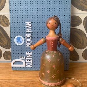 Könnte beinhalten: Eine glockenförmige Holzfigur mit bemaltem Gesicht, braunen Haaren und einem rosafarbenen Oberteil. Der Rock ist olivgrün mit kleinen rosa Blumenmustern und einem rosa Bogenrand. Eine kleine Holzkugel hängt an einer Schnur im Inneren.