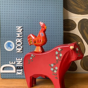 Op de afbeelding: Een rode houten stierfiguur met beschilderde blauwe bloemen en een kleine rode haan erop. De stier heeft kleine witte hoorns. Een blauw bord met de tekst "DE KLEINE NOOR MAN" staat op de achtergrond.