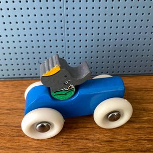 May include: A wooden toy car painted bright blue with four white wheels featuring silver center caps. A gray elephant figure with a yellow crown and a green shirt is attached to the top of the car. The elephant has a small black eye and a trunk.