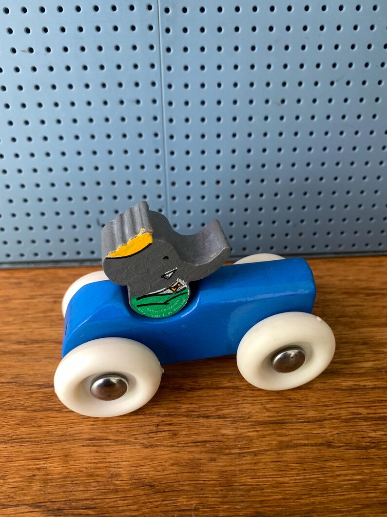 May include: A wooden toy car painted bright blue with four white wheels featuring silver center caps. A gray elephant figure with a yellow crown and a green shirt is attached to the top of the car. The elephant has a small black eye and a trunk.