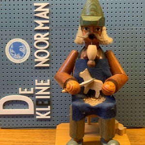 May include: A wooden figurine of a craftsman carving a wooden animal. The figurine wears a green hat, blue apron, and has a long mustache. The text "DE KLEINE NOOR MAN" is visible on the background.