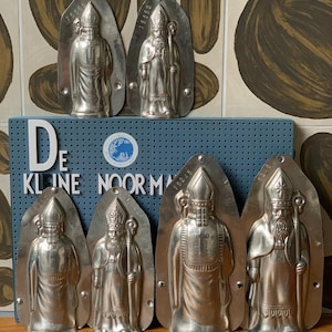 Puede incluir: Colección de moldes de metal plateado vintage con forma de San Nicolás. Los moldes tienen la forma de un obispo sosteniendo un báculo. Los moldes están dispuestos sobre una caja azul con texto blanco.