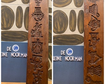 May include: Two vintage wooden cookie molds with intricate carvings. The molds are brown and feature various designs, including a person, a house, and a horse. The molds are labeled "De Kleine Noorman".