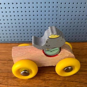 May include: A wooden toy car with a natural light brown finish and four bright yellow wheels. A gray elephant figure with a yellow crown and a green shirt is seated in the center of the car. The wheels are attached with silver metal pins.