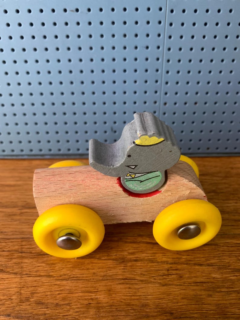 May include: A wooden toy car with a natural light brown finish and four bright yellow wheels. A gray elephant figure with a yellow crown and a green shirt is seated in the center of the car. The wheels are attached with silver metal pins.